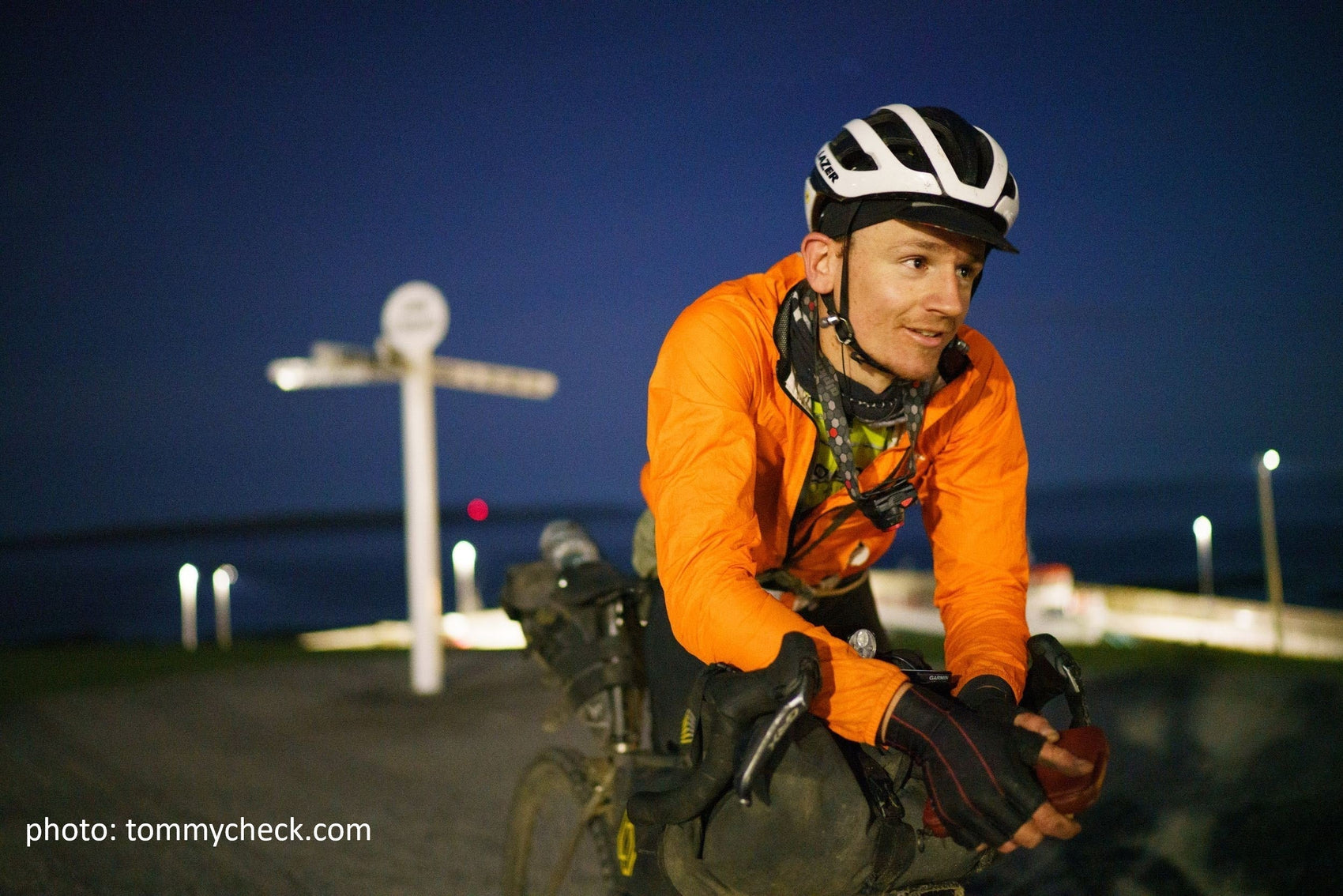 a man in an orange jacket resting on his bike in front of a nightime sky.