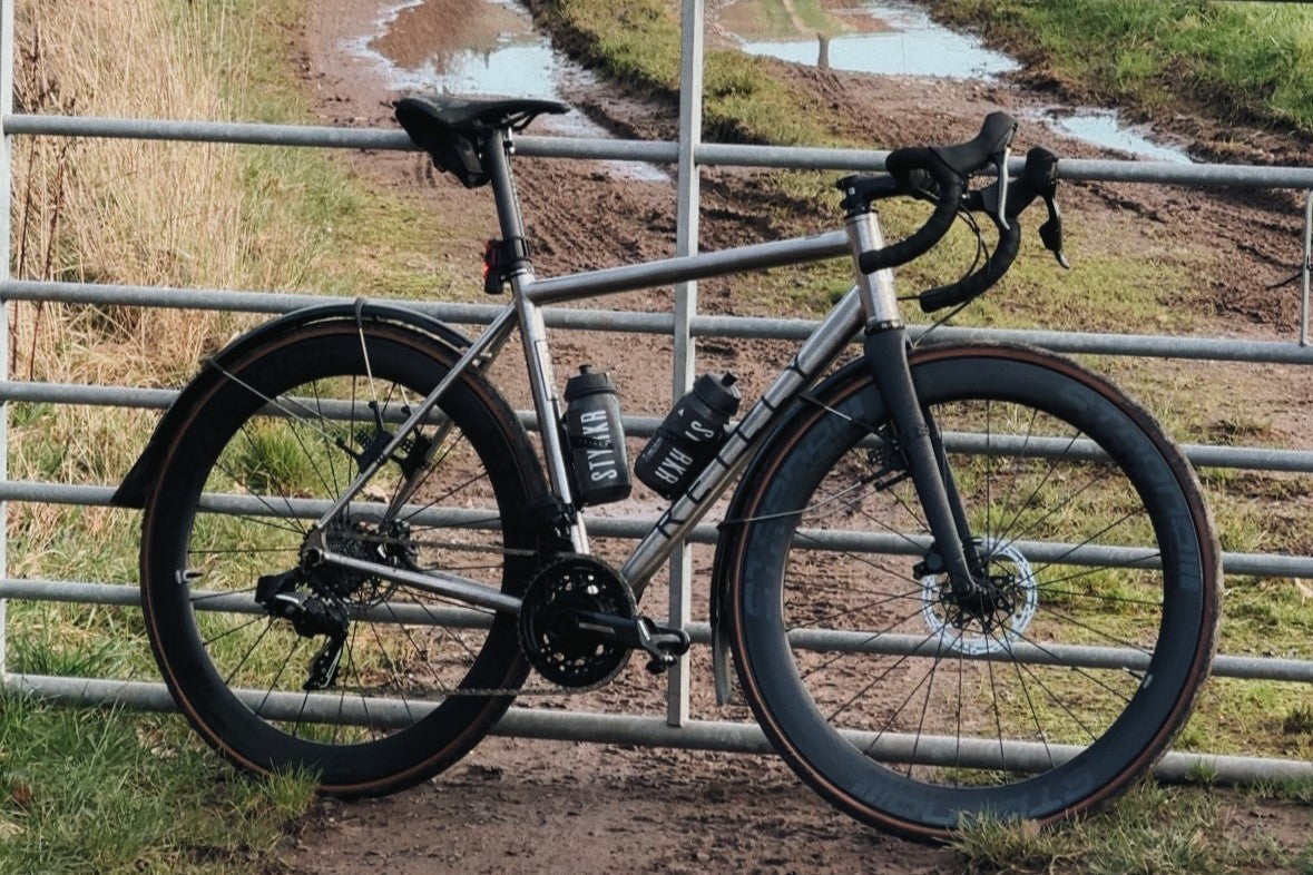 A titanium road bike leans up against a metal gate with muddy field in the background.