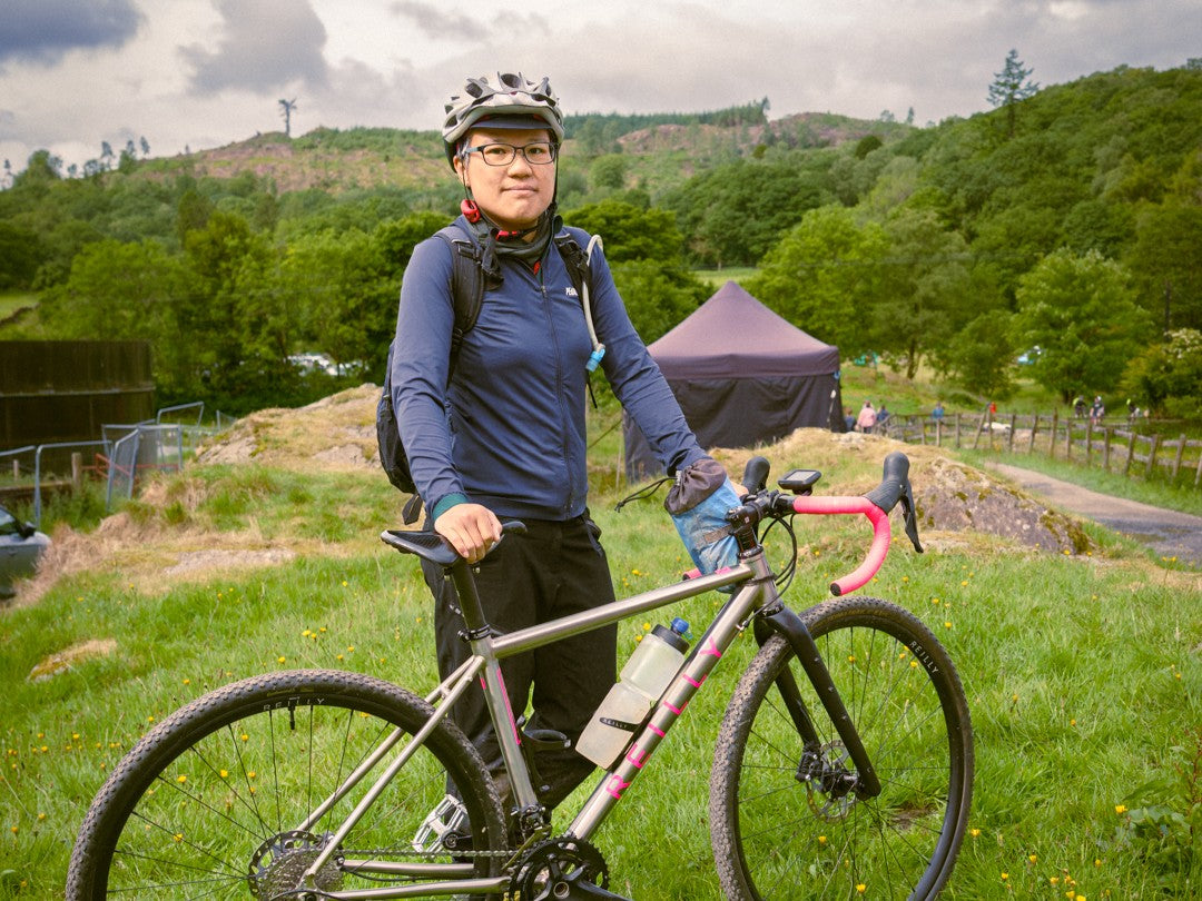 Person with a bicycle in a scenic outdoor setting with greenery and hills.