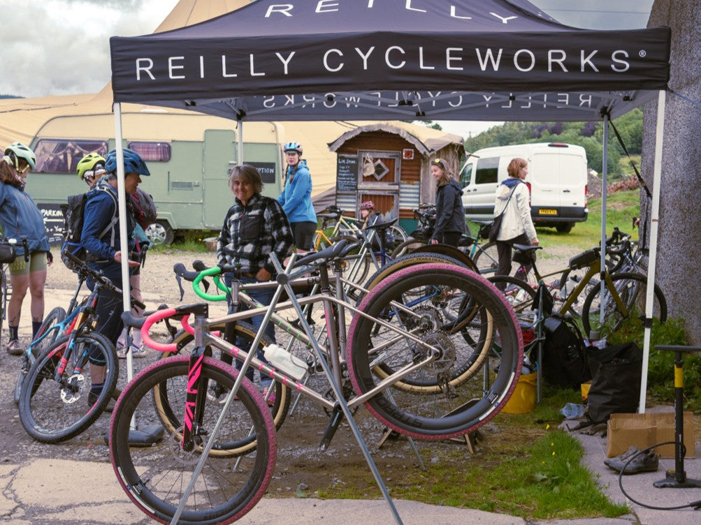 People with bicycles under a Reilly Cycleworks tent at an outdoor event.