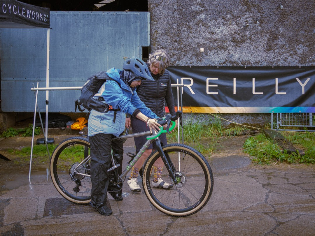 Two people adjusting a bicycle outdoors with 'REILLY' branding in the background.