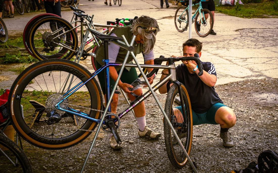 Two cyclists adjusting a bicycle on a road with other bikes and people in the background.