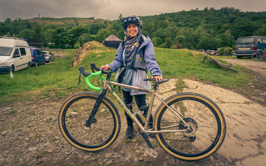 Woman with a bicycle in a rural setting with vehicles and greenery.