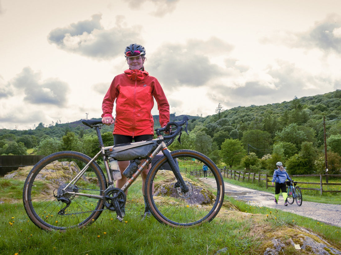 Woman in red jacket standing next to a bicycle on a trail with another cyclist in the background.