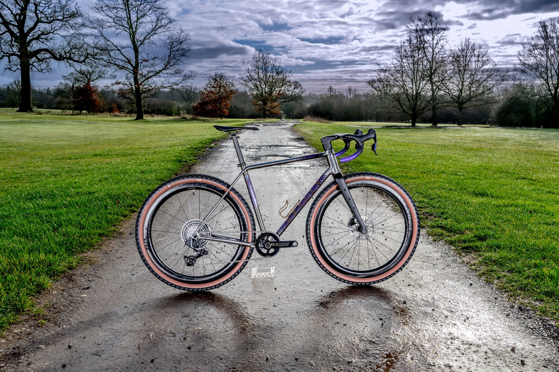 A gravel bicycle with a dark metallic frame and knobby tires stands on a wet dirt path in a grassy park under a cloudy sky.