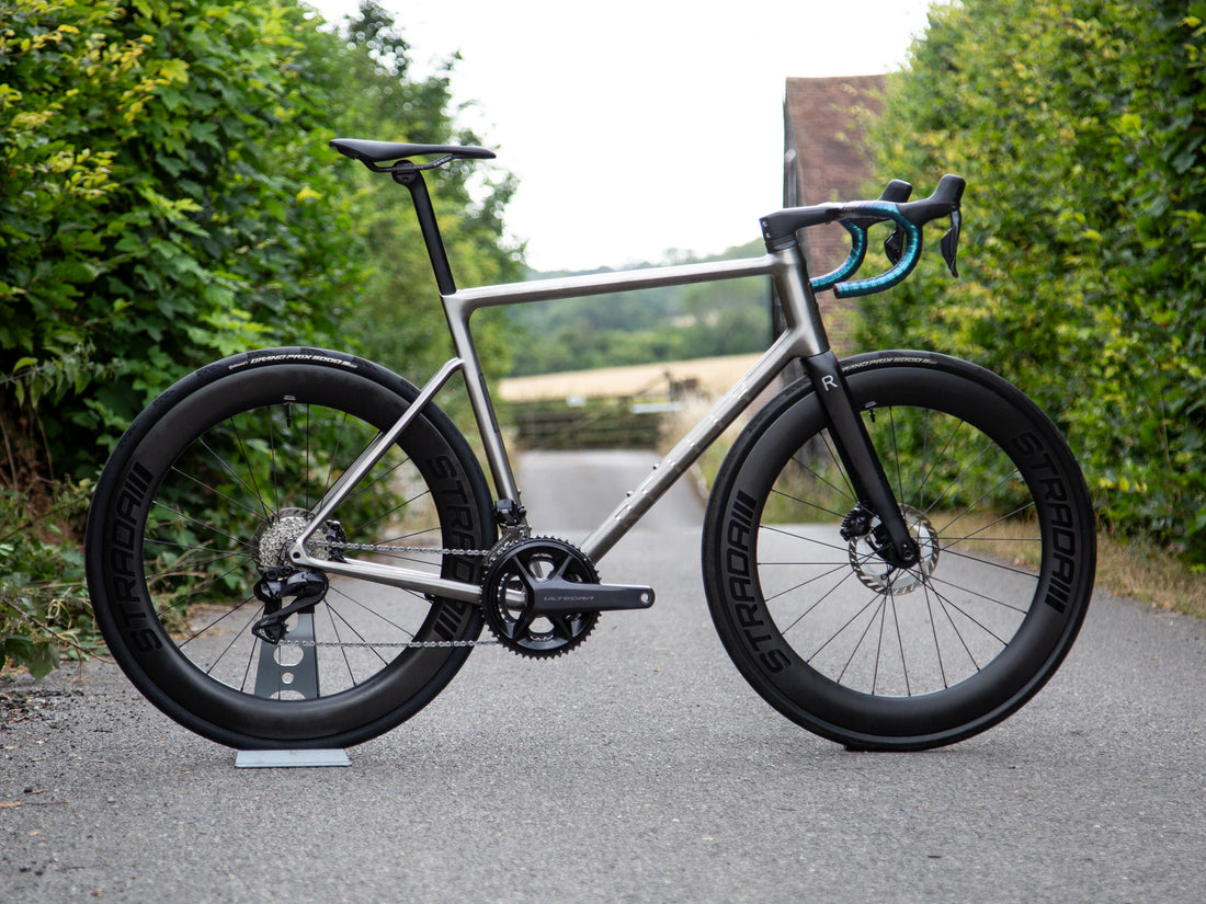 A carbon fiber road/gravel bicycle with black wheels, a black saddle, and blue handlebar tape, photographed outdoors on a paved path with green hedges in the background.