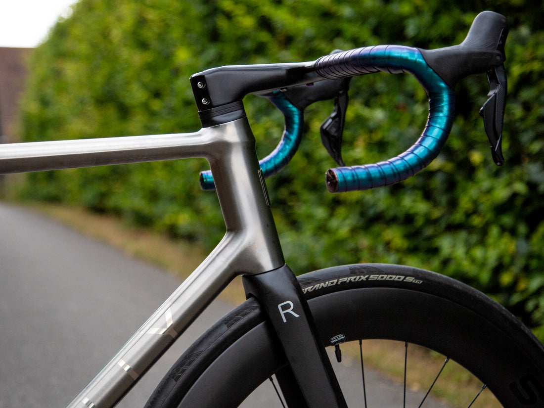 Close-up of a road bicycle frame and drop handlebars with bright blue and black tape wrapping, set against a blurred green hedge and paved path background.