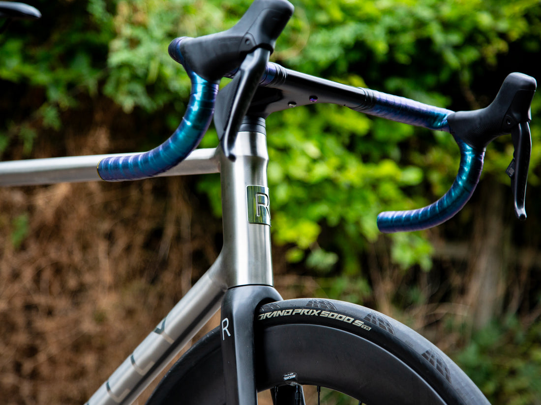 Close-up of a road bicycle handlebar area with black drop handlebars, blue tape wrapping, a silver frame tube, and a black tire against a blurred green outdoor background.