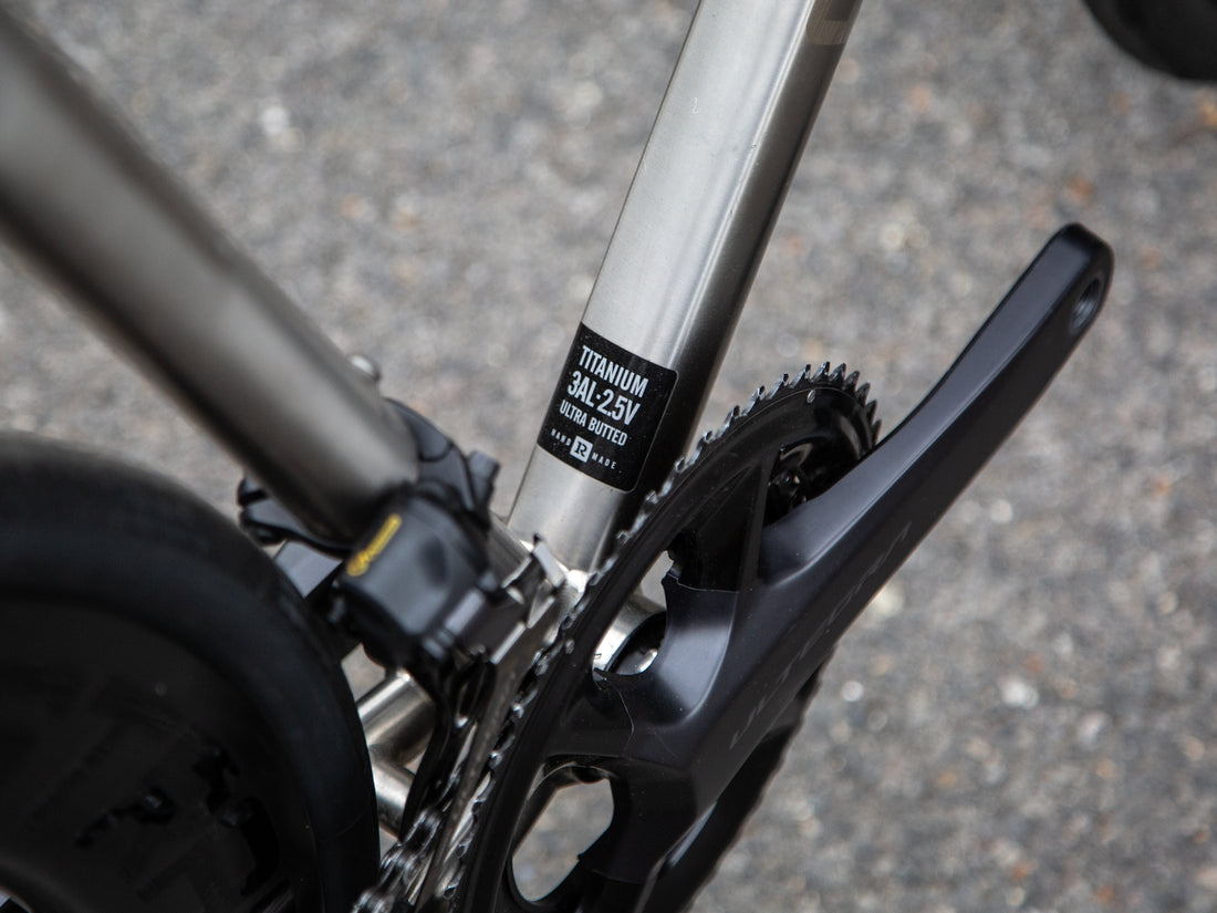 Close-up of a bicycle frame showing a black label that reads 'TITANIUM 3AL-250' and 'SILVER BIFERRO', with part of a black crankset and tire visible against a blurred gray pavement background.