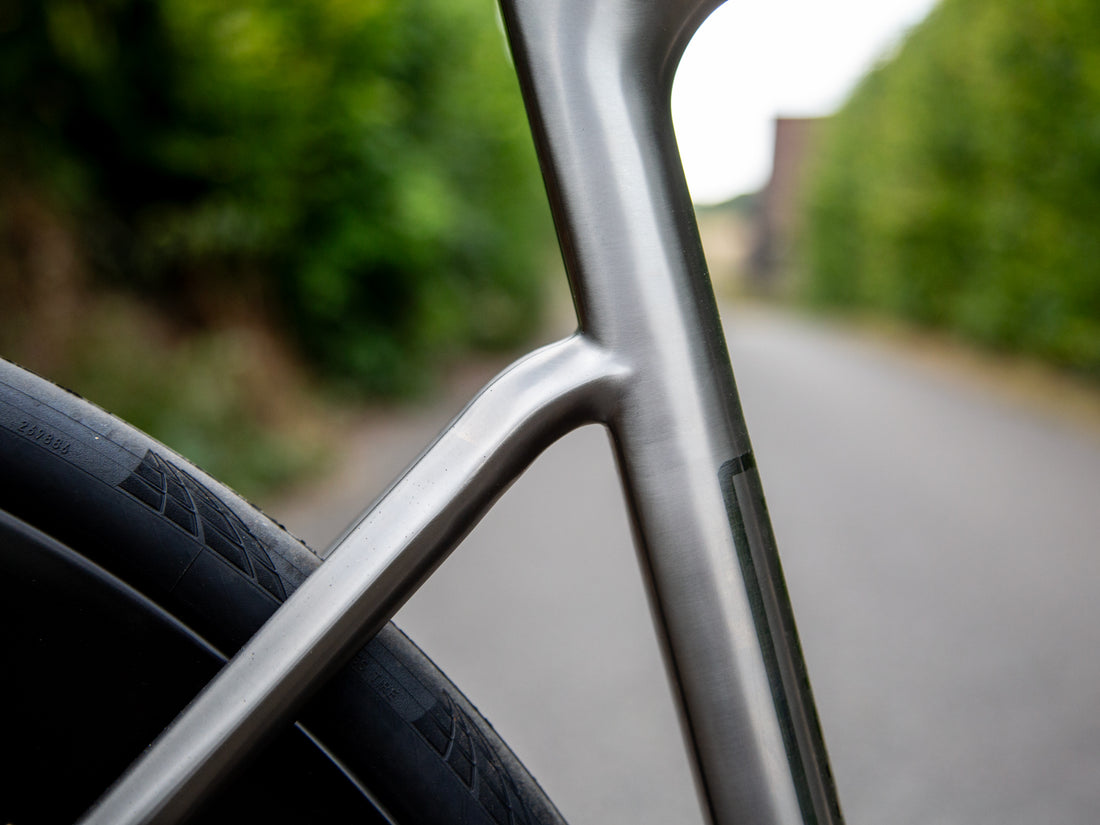 Close-up of a bicycle frame and front wheel with a black tire, photographed outdoors on a blurred road and greenery background.