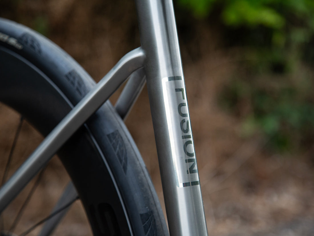 Close-up of a bicycle frame with the word 'FUSION' printed vertically on a metallic silver tube, with part of a black tire visible against a blurred outdoor background.
