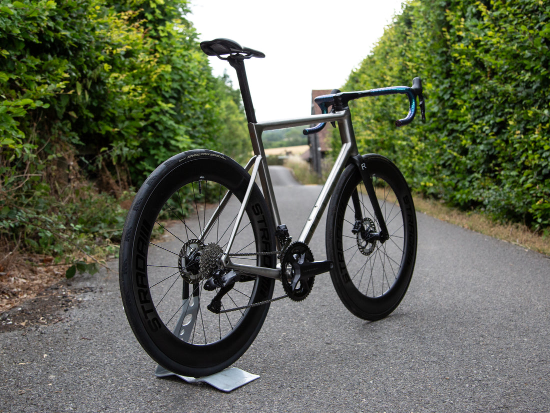 A carbon fiber road bicycle with black wheels and blue handlebar tape, photographed outdoors on a paved path between green hedges.