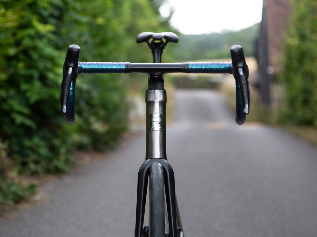 Close-up front view of a drop-bar bicycle handlebar and frame on a blurred country road with green foliage on both sides.