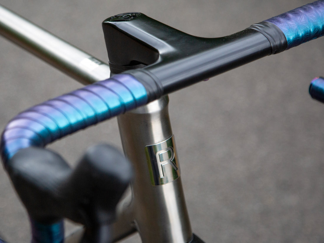 Close-up of a bicycle handlebar area showing a black stem, silver frame tube with a metallic 'R' emblem, and drop handlebars wrapped in iridescent blue-purple tape against a blurred gray background.