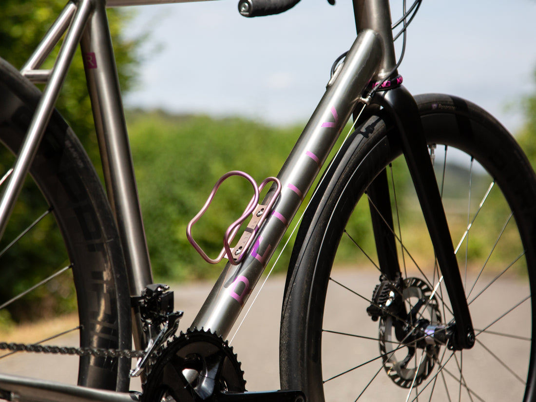 Close-up of a titanium bicycle frame with pink accents, a pink water bottle cage, and black wheels, photographed outdoors on a blurred road and greenery background.