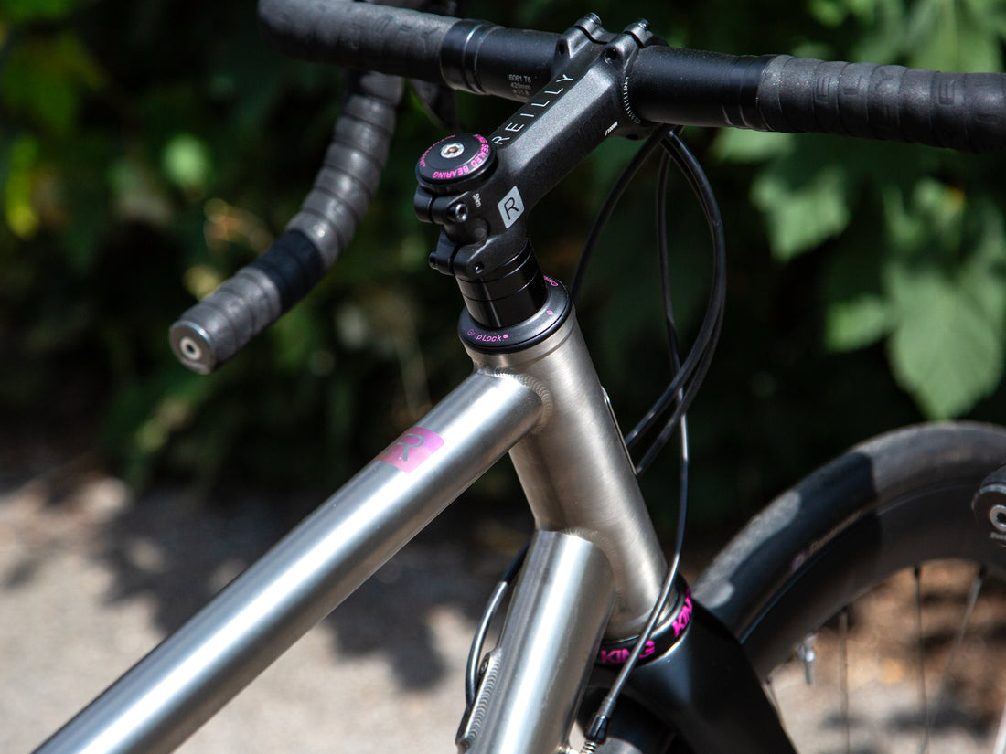 Close-up of a bicycle handlebar and frame area, showing a matte black stem, black handlebar tape, black fork, and a titanium frame with small pink accents, photographed outdoors with blurred green foliage in the background.