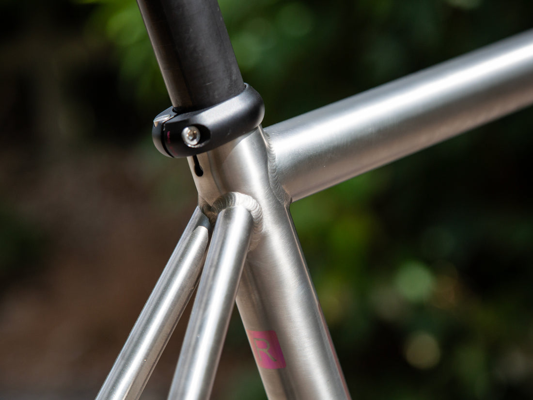 Close-up of a silver metallic bicycle frame with a black seat post clamp and a small pink logo near the bottom tube, set against a blurred green outdoor background.