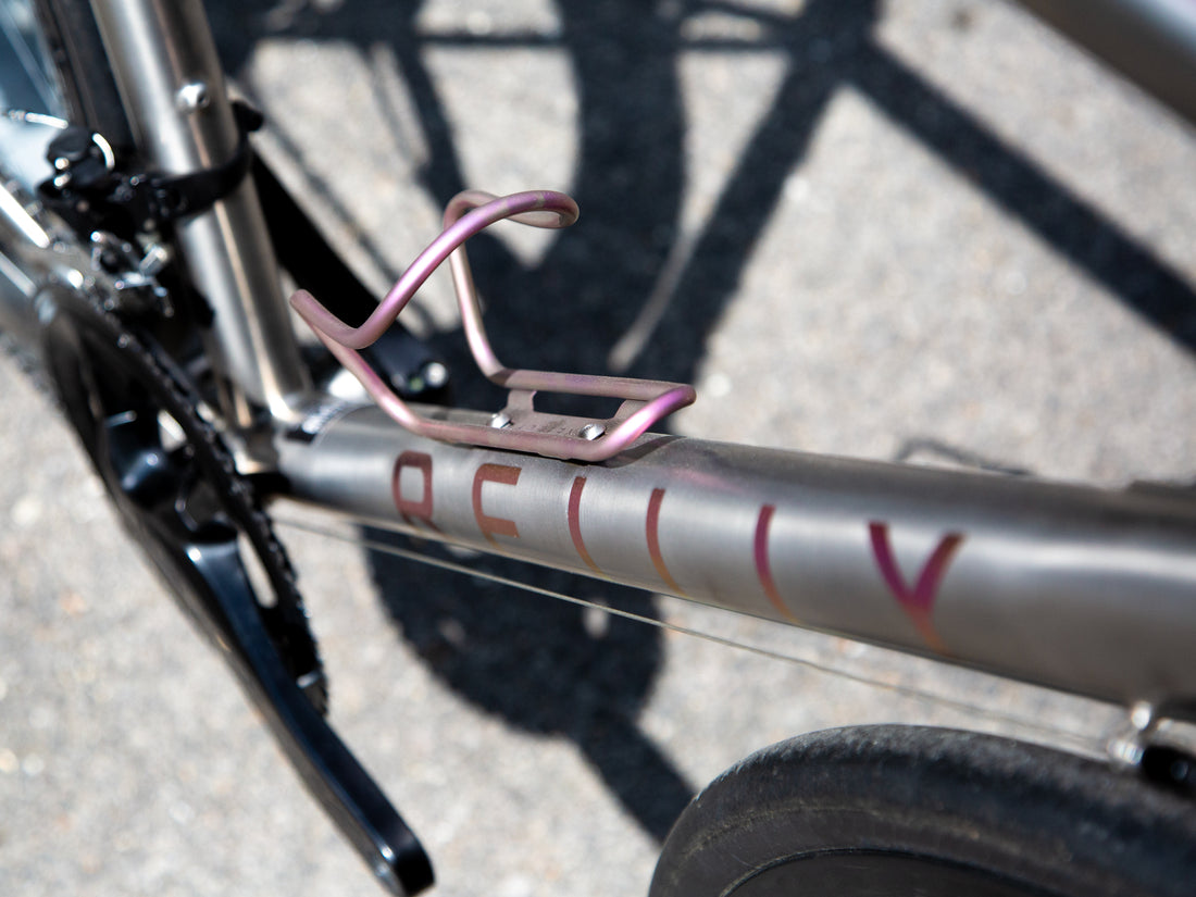 Close-up of a bicycle frame with a metallic rose gold water bottle cage mounted on the down tube. The frame has 'REILLY' printed on it in rose gold lettering, and the background shows asphalt with bicycle wheel shadows.