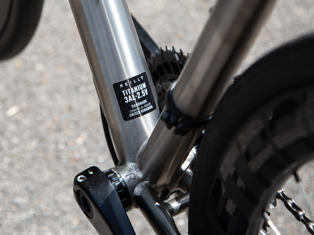 Close-up of a bicycle frame showing a silver titanium down tube with a black label that reads 'REILLY TITANIUM 3A+2.5V' 
