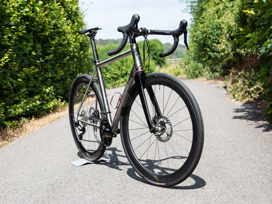 A titanium bicycle with black components and drop handlebars, parked on an asphalt path outdoors with green hedges on both sides.