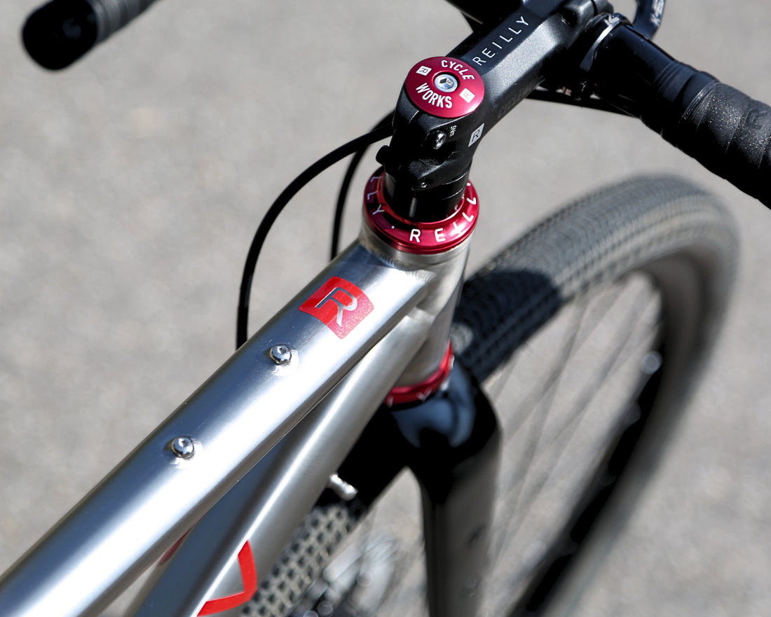 Close-up of a bicycle frame and handlebar area showing a silver aluminum frame with red accents, a black stem, and a red RockShox Pike fork end on an outdoor paved surface.