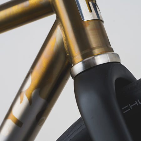 Close-up detail of a bicycle frame with a metallic gold and silver finish, showing the seat tube junction and part of a black fork.