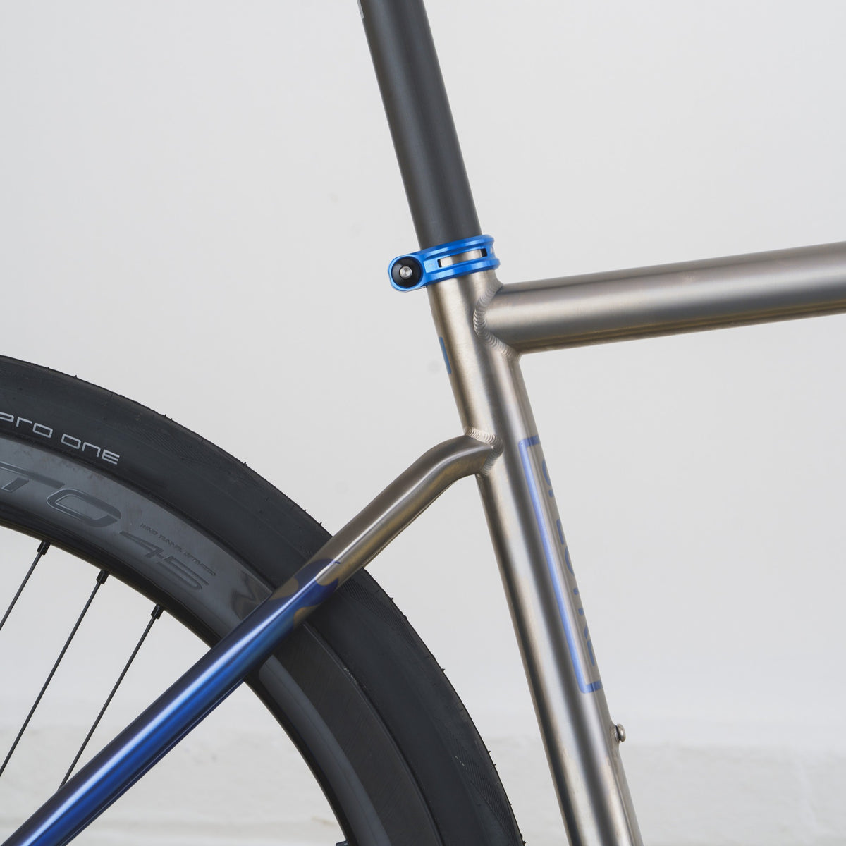 Close-up of a bicycle frame showing a metallic silver down tube, blue seat-tube clamp, and blue down-bracket over a black tire against a light gray background.