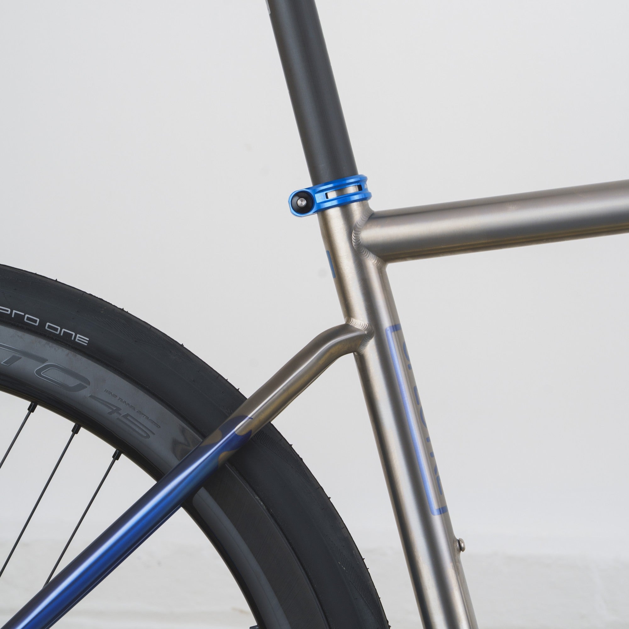 Close-up of a bicycle frame showing a metallic silver down tube, blue seat-tube clamp, and blue down-bracket over a black tire against a light gray background.