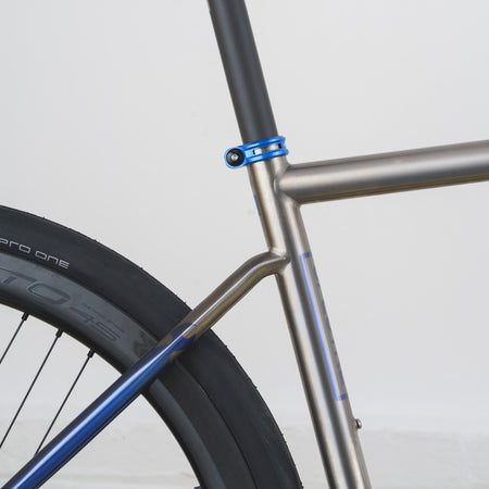 Close-up of a bicycle frame showing a metallic silver down tube, blue seat-tube clamp, and blue down-bracket over a black tire against a light gray background.