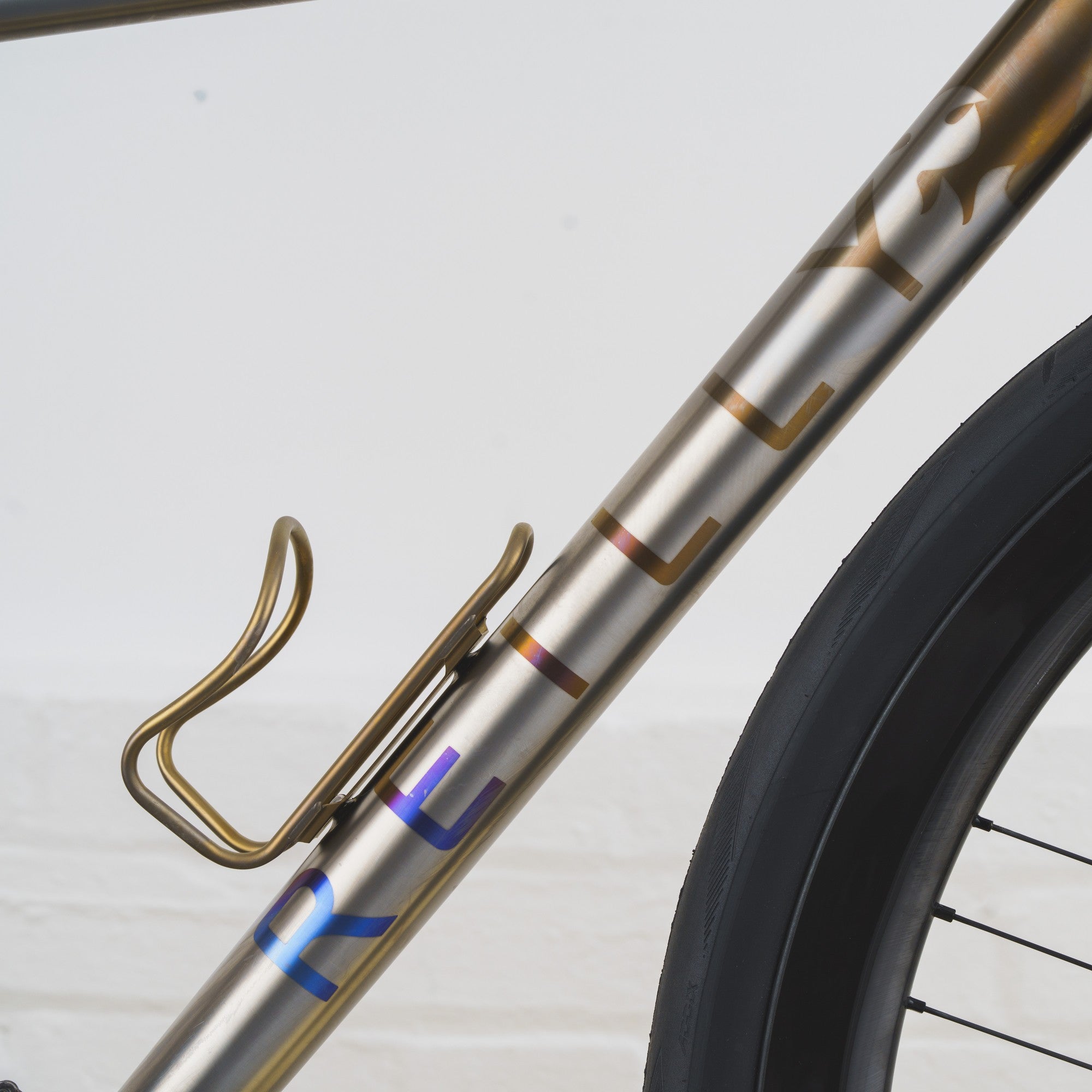 Close-up of a bicycle frame with a gold-finished metal finish, a gold water bottle cage, and partial iridescent lettering near the bottom, alongside a black tire against a light gray background.