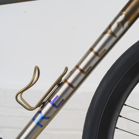 Close-up of a bicycle frame with a gold-finished metal finish, a gold water bottle cage, and partial iridescent lettering near the bottom, alongside a black tire against a light gray background.