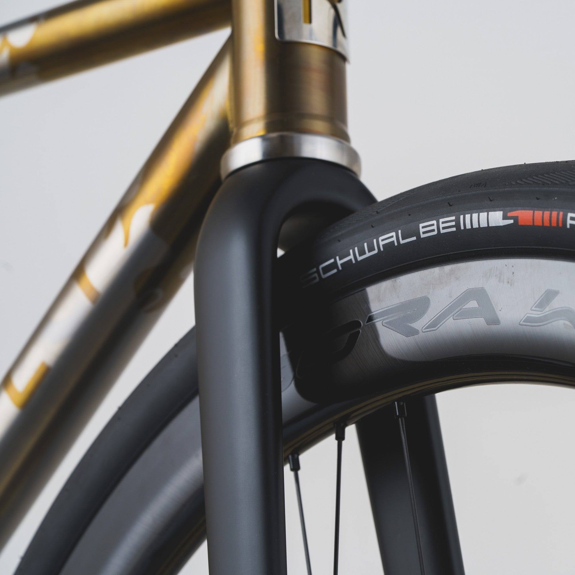 Close-up of a bicycle wheel and frame showing a black Schwalbe GT radial tire mounted on a dark rim, with a metallic gold and silver bicycle frame in the background.