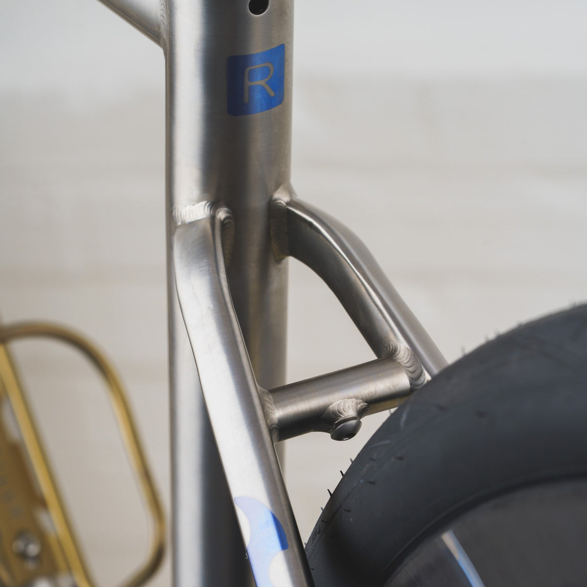 Close-up of a metallic bicycle frame with a blue logo featuring a stylized letter 'R' and part of a black tire visible on the right.