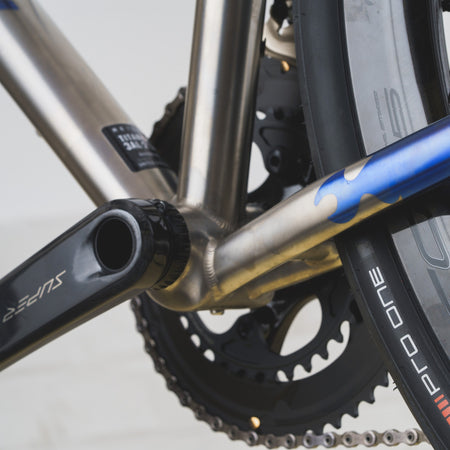 Close-up of a bicycle frame and crankset showing a metallic silver frame tube, a black Shimano crank arm, a blue accent on the down tube, and part of a black Pro One tire.