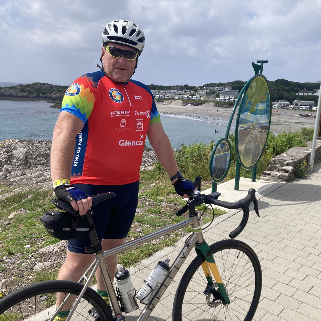 a man in a red cycle shirt, holding his bike in front of the sea.