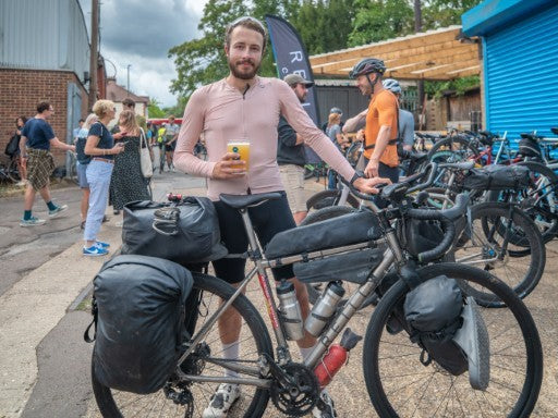 Man with a bicycle loaded with gear in an outdoor setting holding a beer