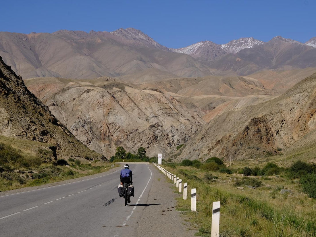 A cyclist rides along a road with snow capped mountains in the foreground.