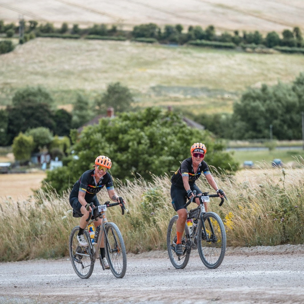 Two cyclists riding on a gravel road with a scenic background of fields and trees.