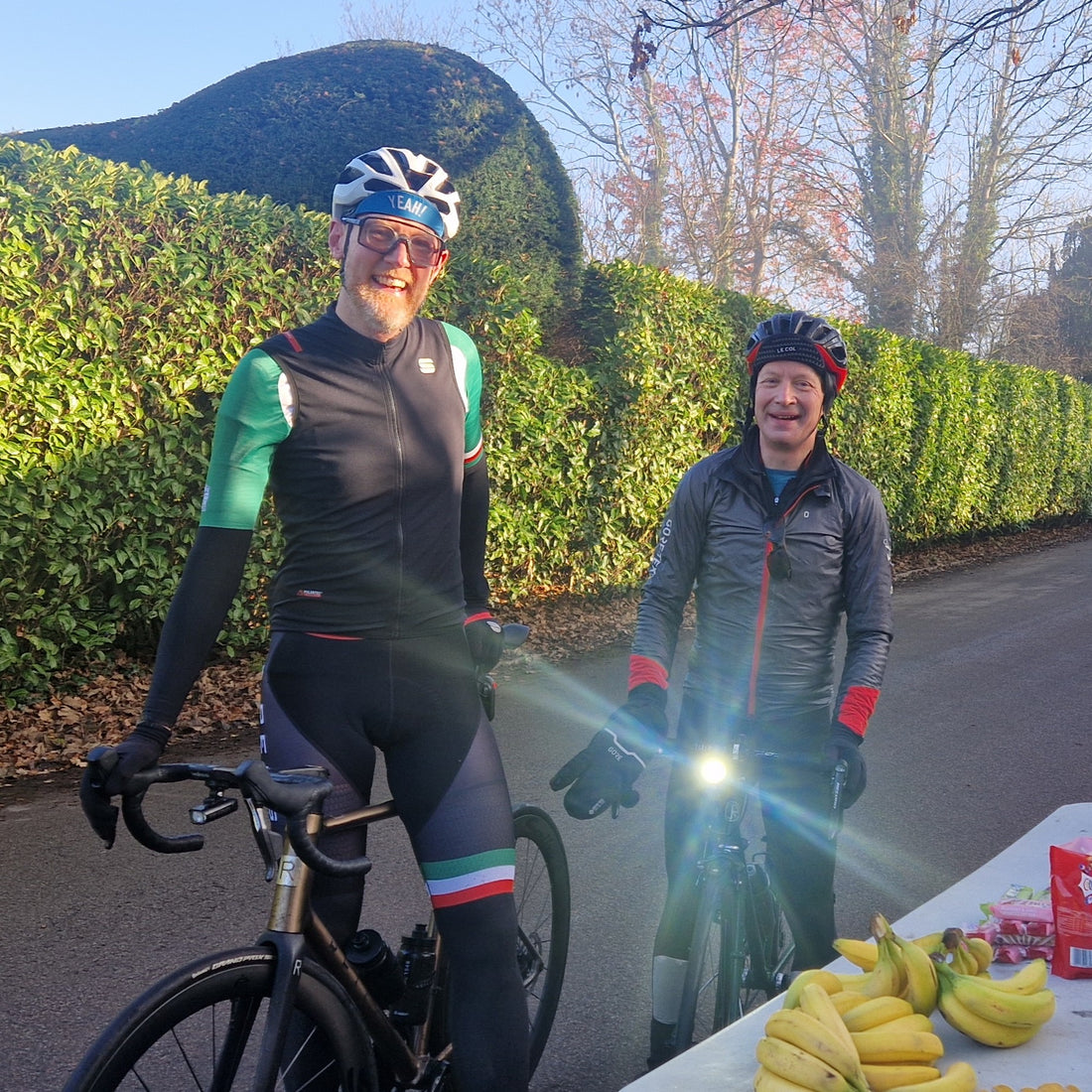 Two cyclists standing on a road with a table of bananas and other items in the foreground.