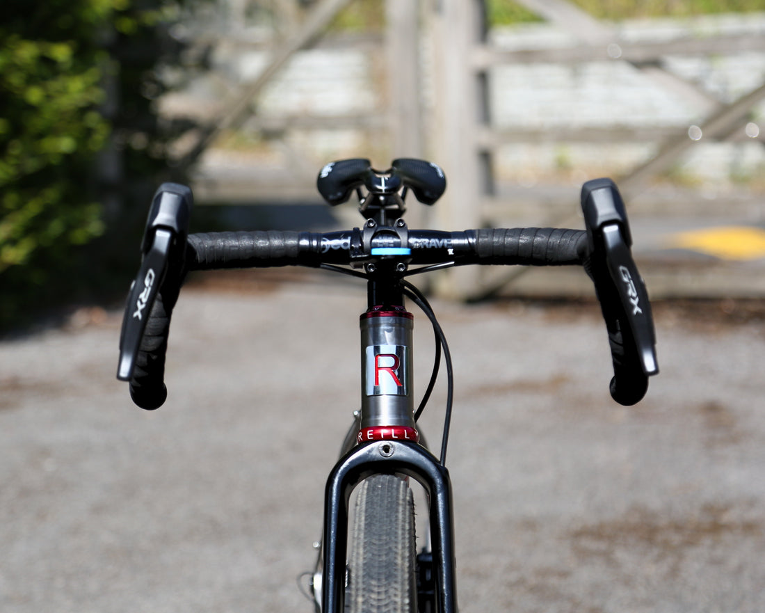 Close-up front view of a road bicycle handlebar and fork, with black drop handlebars, black brake levers branded XDR, and a dark gray carbon frame with a red and white R1 brand decal, set against a blurred outdoor background.