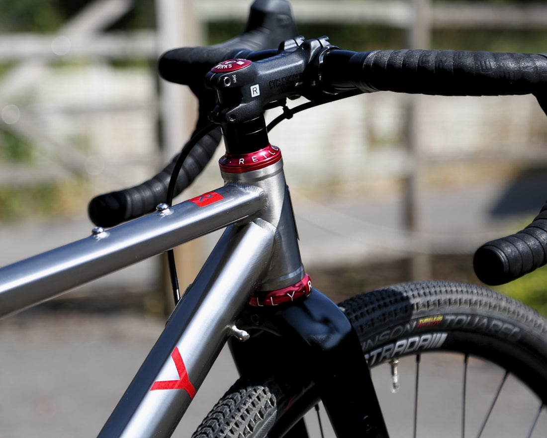 Close-up of a bicycle frame and handlebar area with a silver metallic frame, black drop handlebars, black fork, and red accent rings on a blurred outdoor background.
