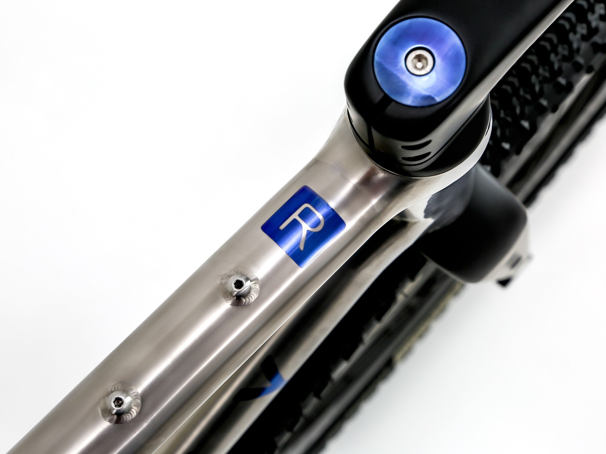 Close-up of a bicycle frame showing a brushed silver aluminum tube with a blue square logo containing a white letter 'R', alongside black components and a blue seat post clamp on a white background.