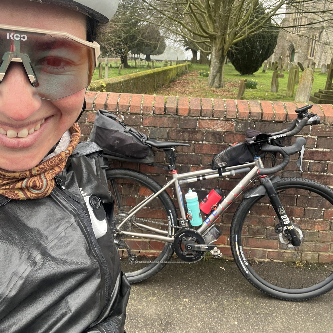 A close-up selfie of a smiling cyclist wearing a helmet and KGO glasses, standing next to a silver gravel bicycle loaded with black bags, parked against a brick wall in a cemetery.