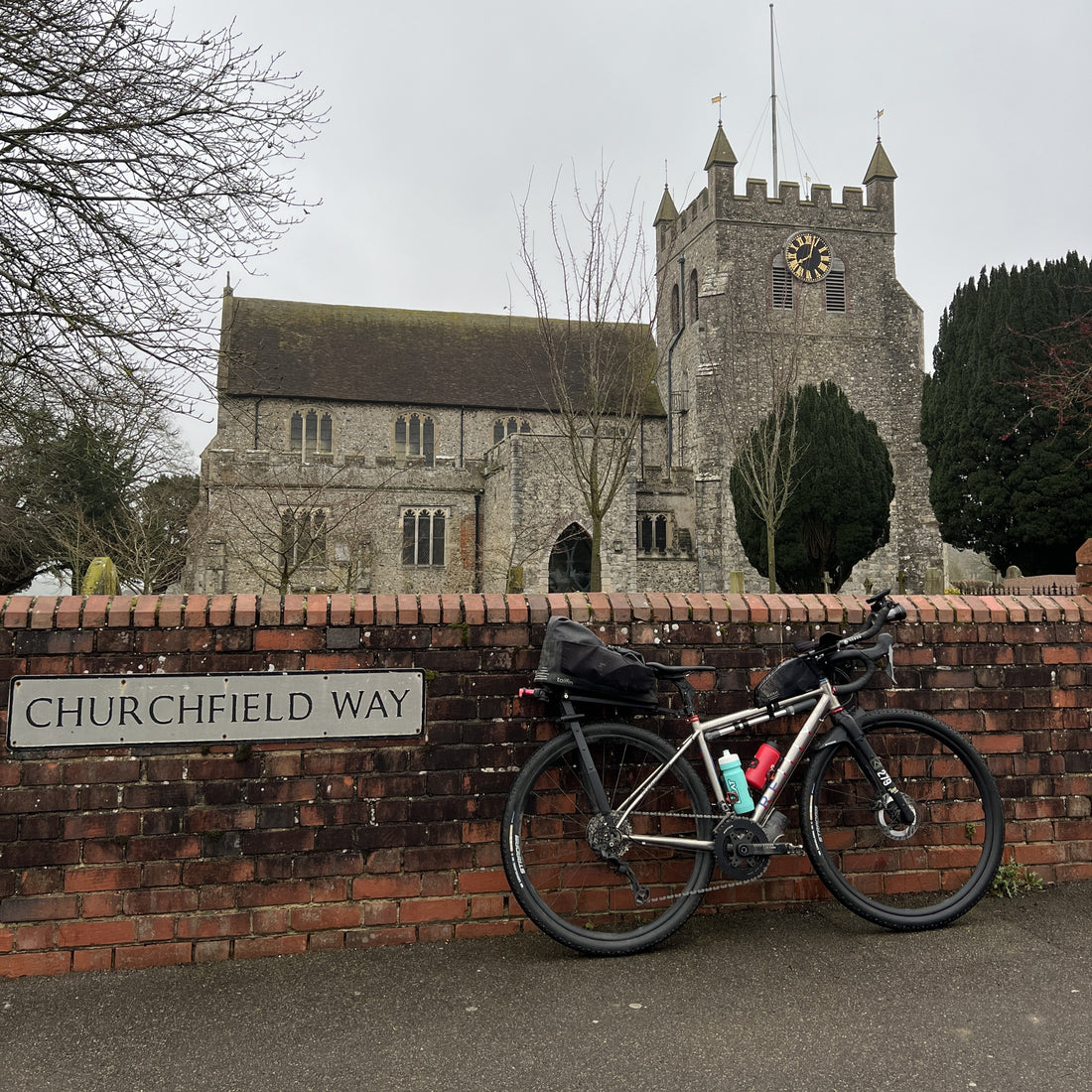 A black road bicycle with rear and front racks, a top tube bag, and water bottles is parked against a brick wall next to a street sign reading 'CHURCHFIELD WAY'. In the background is a stone church tower and building under an overcast sky.