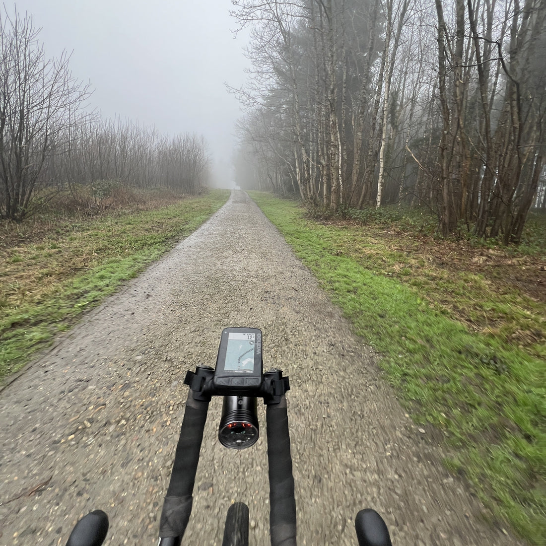 A first-person view from a cyclist riding on a gravel path through a foggy forest, with a bike computer mounted on the handlebars in the foreground.
