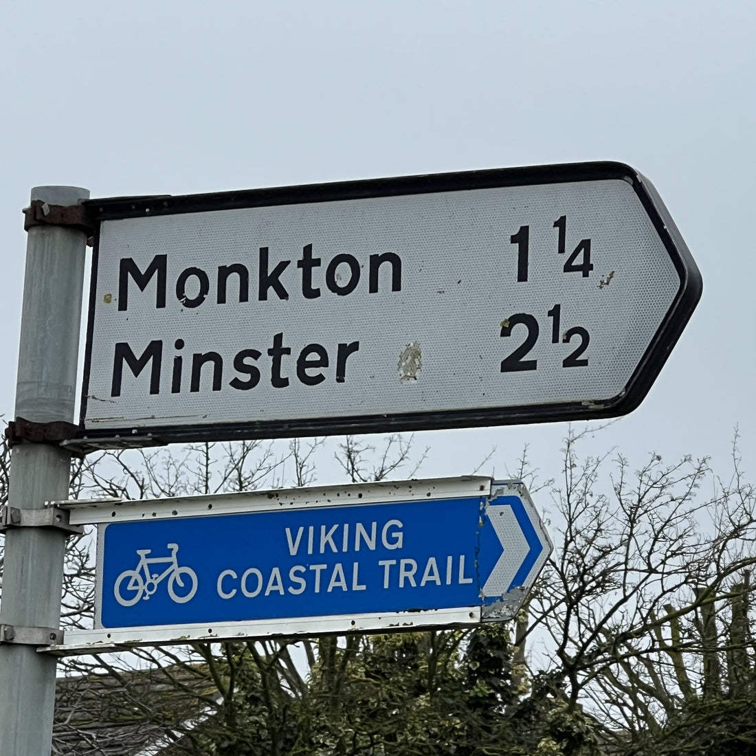 A real-world outdoor scene showing two directional road signs on a metal post against a pale sky and bare trees. The top sign reads 'Monkton 1 1/4' and 'Minster 2 1/2' in black text on a white arrowboard.