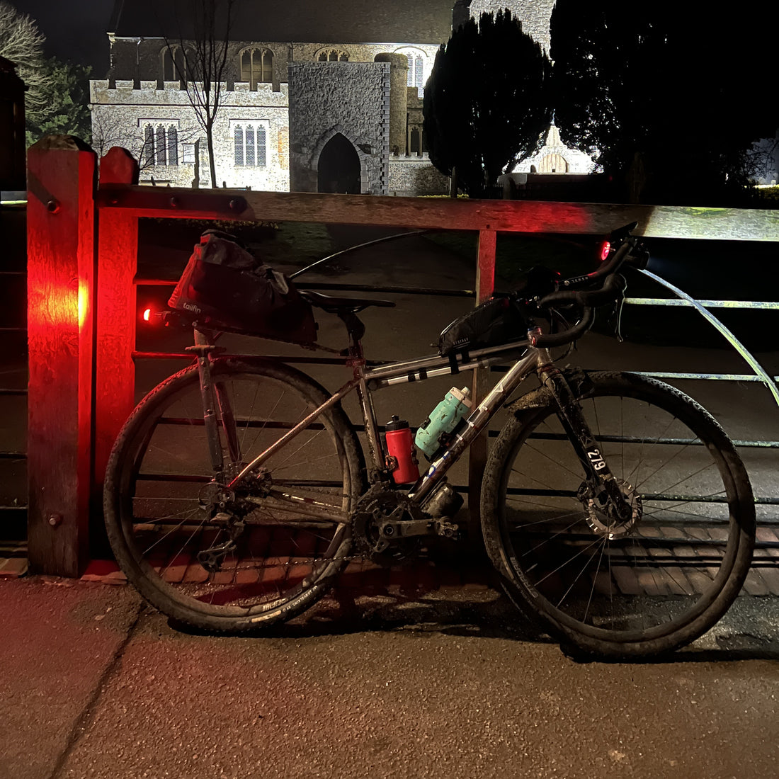 A full-suspension gravel bicycle with rear and front racks, a top tube bag, and water bottles, parked against a wooden bridge railing at night with a stone building illuminated in the background.