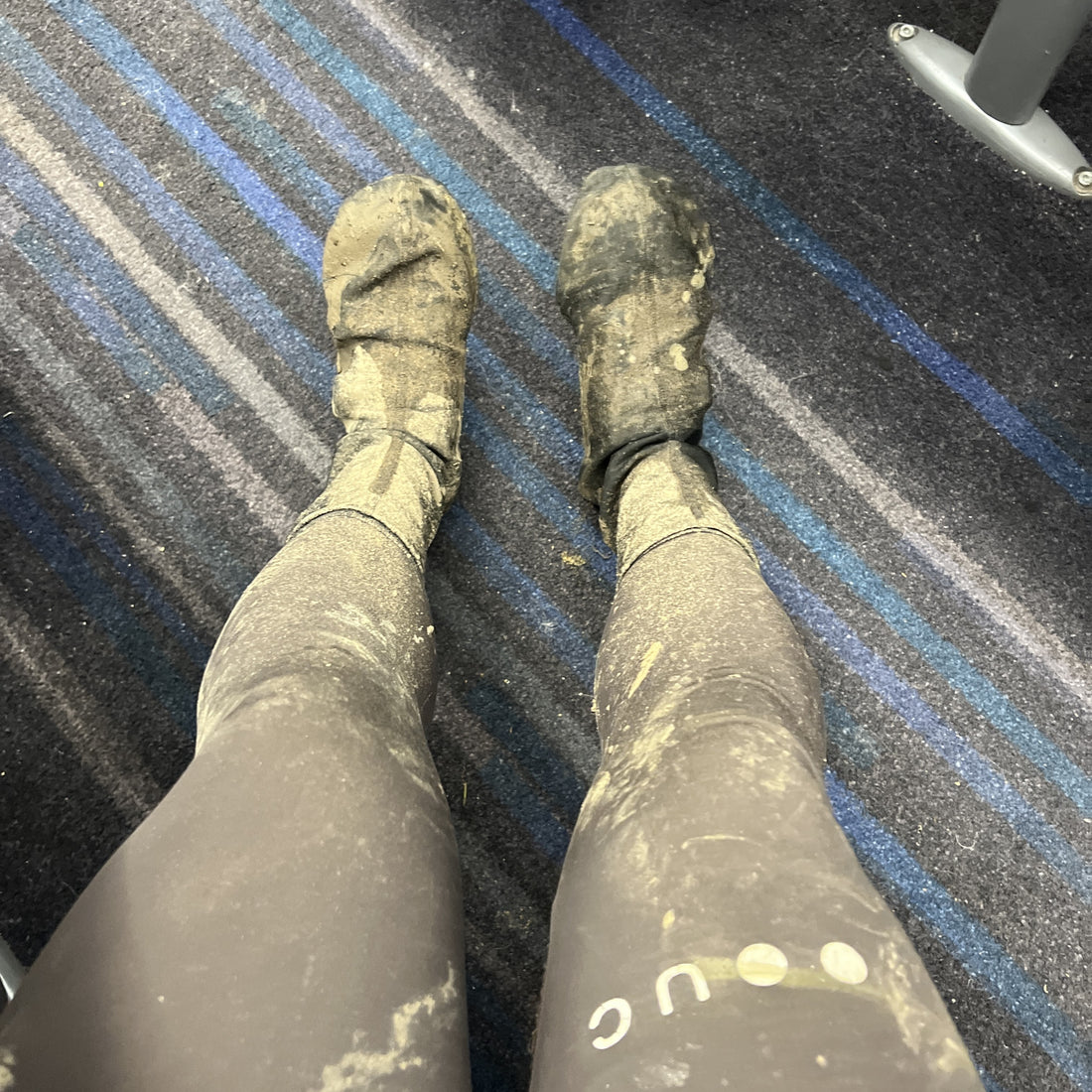 A first-person view of legs wearing dirty, dusty black athletic leggings and boot covers standing on a blue and gray striped carpet.