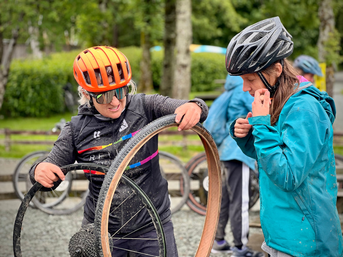 Two people with bicycle helmets, one fixing a wheel, in an outdoor setting with trees and other people in the background.