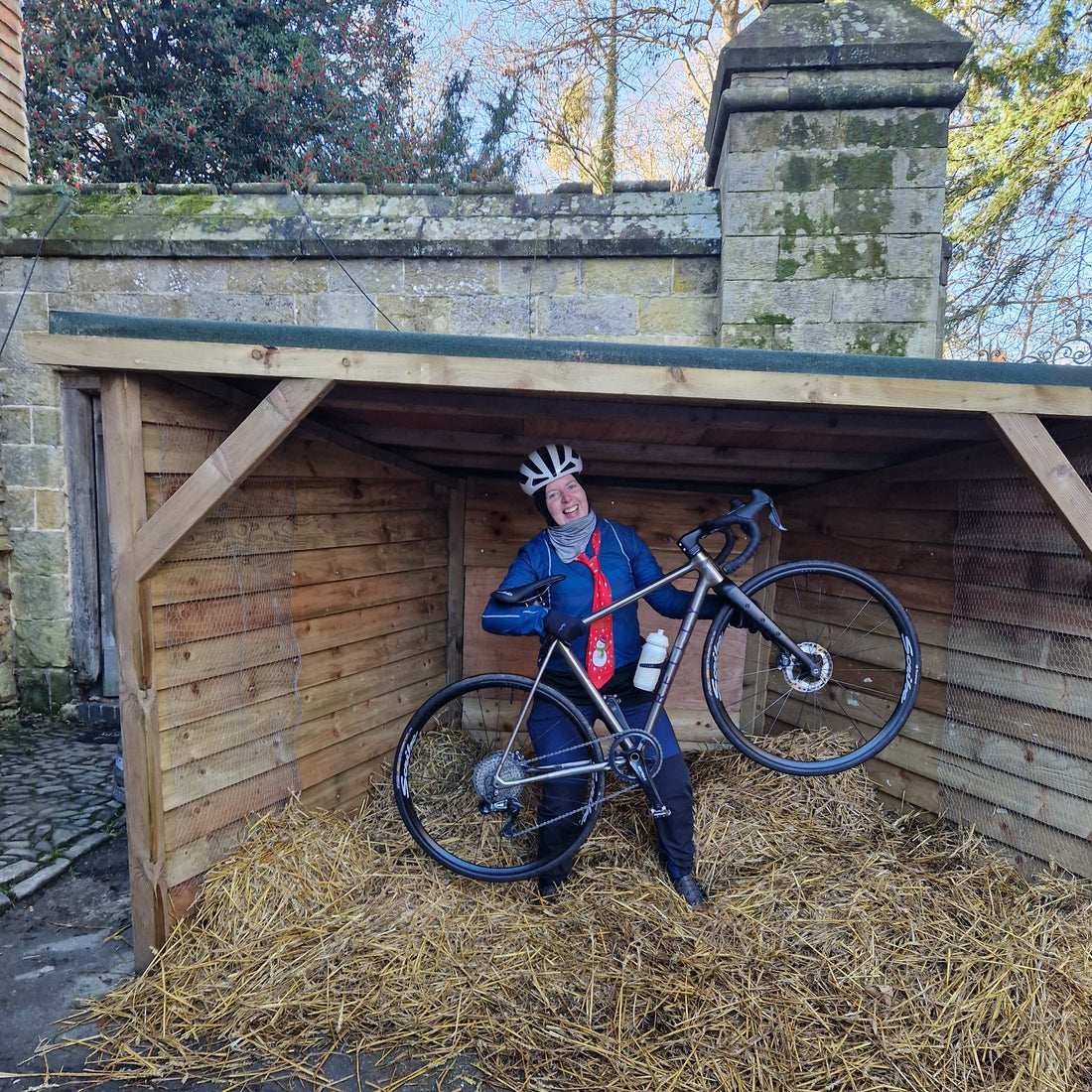 Person with a bicycle standing under a wooden shelter with hay on the ground.